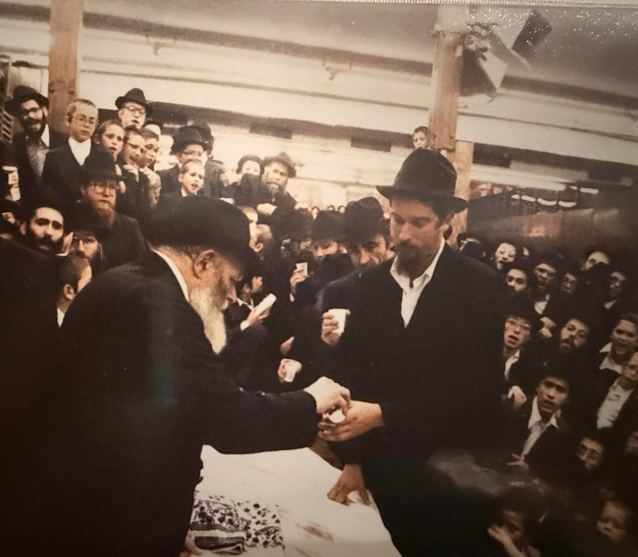 En stor forsamling av mennesker omgir en mann som sitter ved et bord og utveksler noe med en annen mann. Mange har på seg hatter og dresser