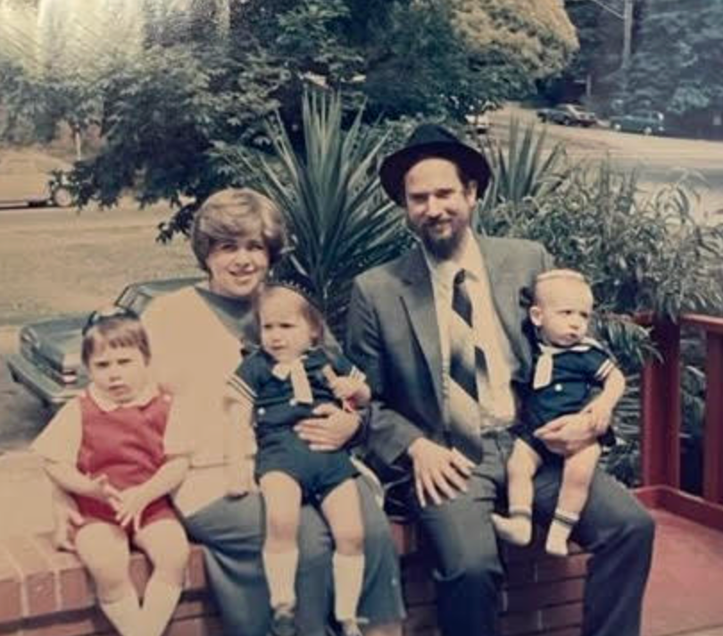 En familie sitter utendørs på en murvegg. To voksne med hatter sitter med tre små barn i matchende antrekk. Trær og biler er synlige i bakgrunnen