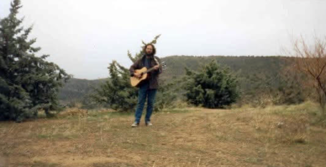 Person som står utendørs med en gitar, omgitt av trær og åser i bakgrunnen, og skaper en rolig, naturlig setting