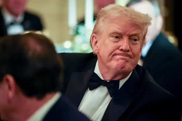 En person i smoking på et arrangement, ser til siden med et nøytralt uttrykk. Bakgrunnen har formell middagssetting