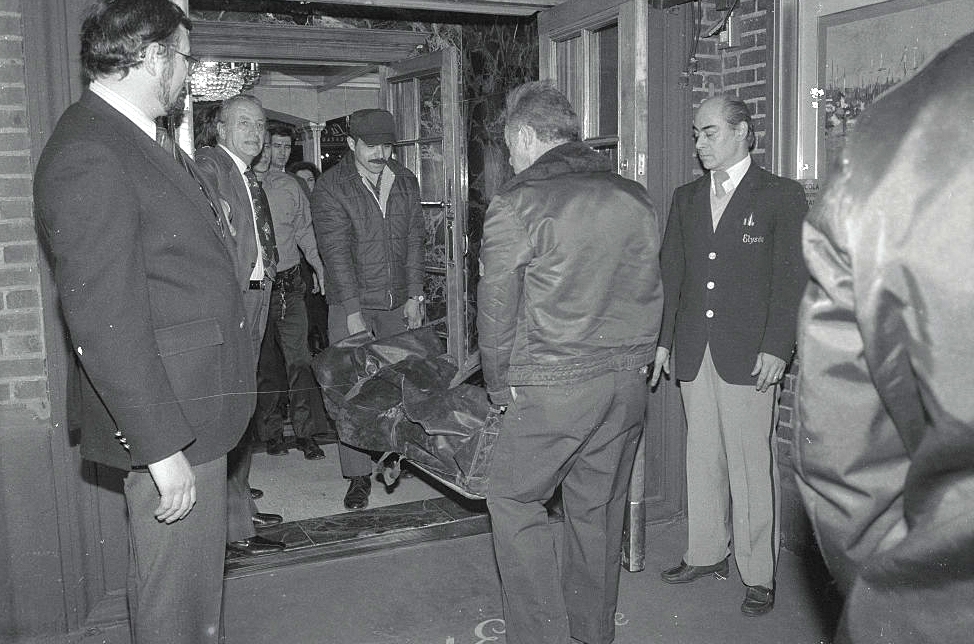 Gruppe menn i dress og jakker som står rundt mens tjenestemenn bærer en båre med en tildekket figur gjennom en døråpning