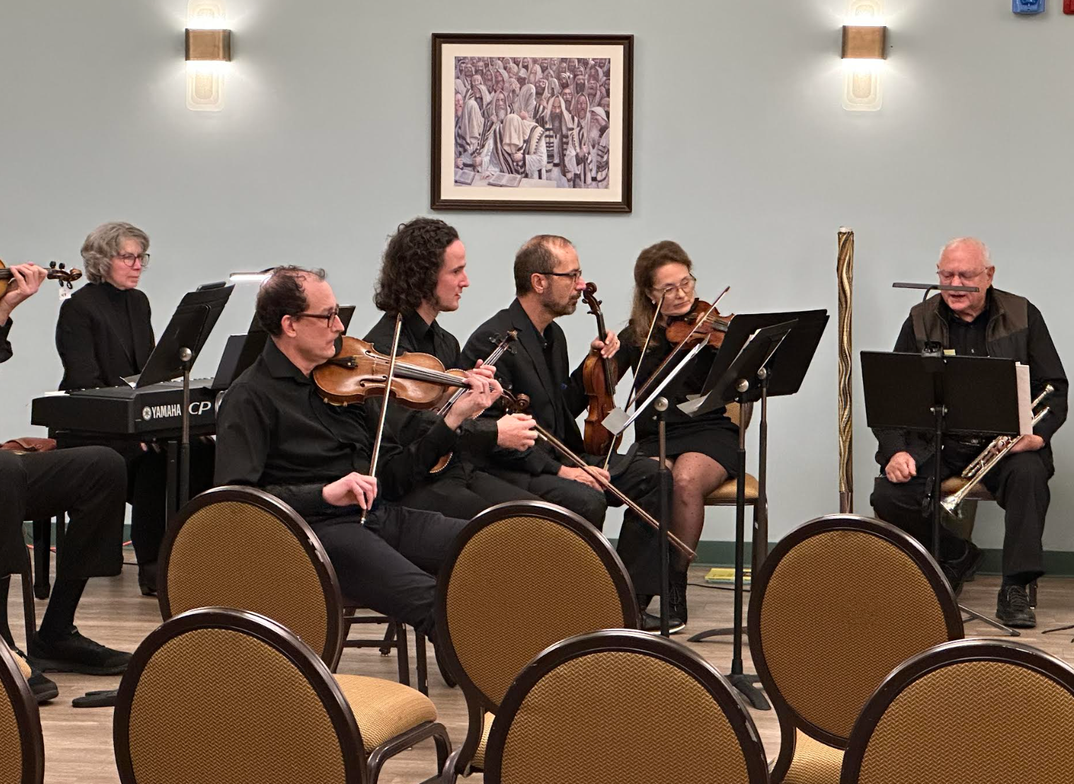 Musikere i formell antrekk opptrer i et lite ensemble med fioliner, en trompet og et keyboard i et rom med innrammet kunstverk på veggen
