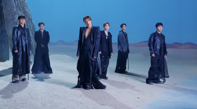 Seks personer i futuristiske, flytende antrekk står på et ørkenlignende sett med en steinete bakgrunn