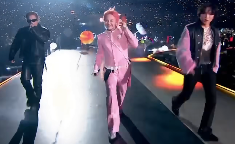 Tre utøvere på scenen; en i et svart skinnantrekk, en i en rosa dress og en annen i en stilig jakke med grafisk t-skjorte som gir energi til publikum