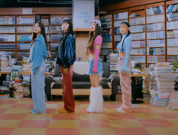 Fire personer står i et bibliotek med hyller og bøker, som hver viser særegen mote, inkludert denim, skinn og en blanding av uformelle stiler