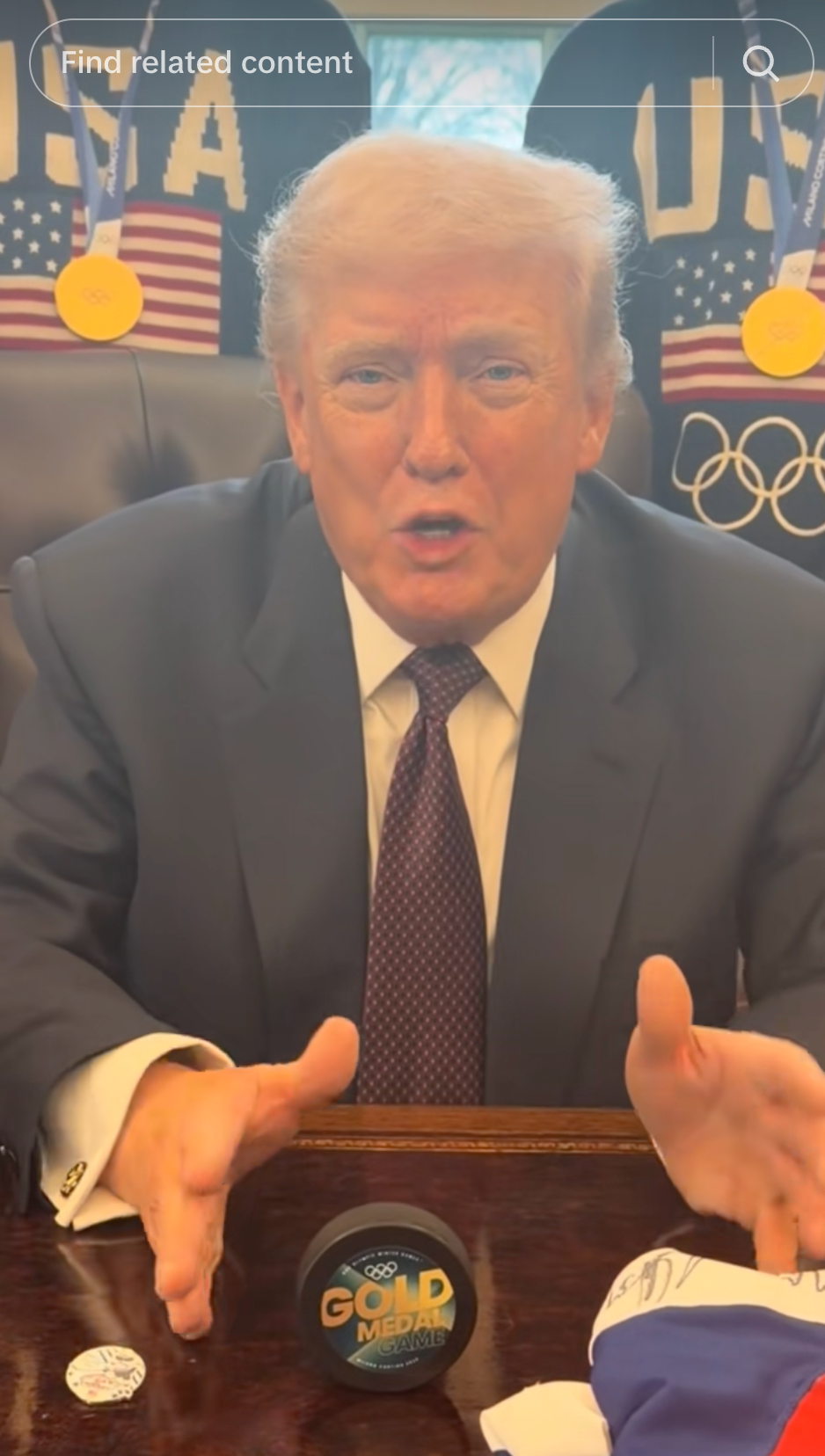 En person i dress sitter ved et skrivebord med gjenstander med olympisk tema, inkludert en gullmedaljeutstilling, og snakker med uttrykksfulle håndbevegelser