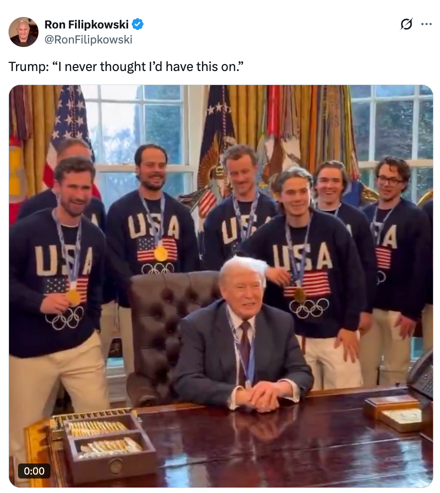 En gruppe menn iført USA OL-gensere poserer med en sittende mann i dress og slips ved et trepult inne i et formelt rom