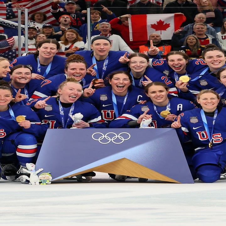 Gruppe kvinnelige idrettsutøvere i ishockeydrakter feirer på banen med olympiske medaljer, omgitt av jublende fans og flagg i bakgrunnen