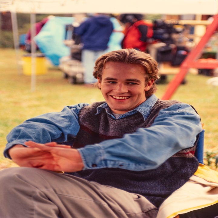 Smilende person sitter utendørs, iført en uformell denimskjorte og genservest, med filmutstyr synlig i bakgrunnen