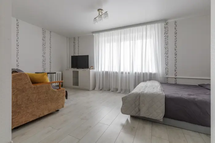 Minimalistisk soverom med seng, TV på stativ og brun sofa. Rene gardiner dekker et stort vindu og slipper inn naturlig lys