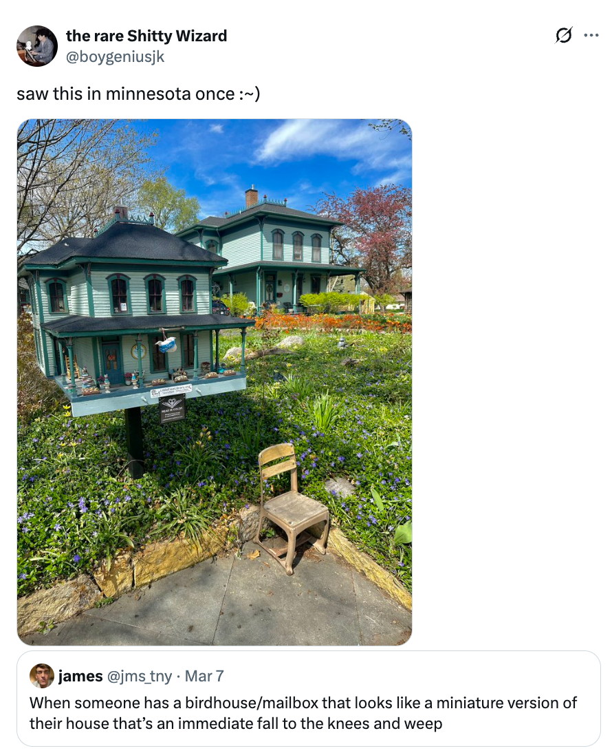 Et stort fuglehus som ligner en miniatyrversjon av et nærliggende hus i historisk stil, omgitt av en hage med en liten stol foran