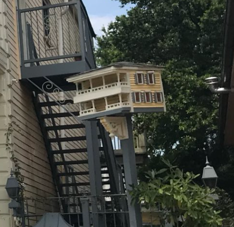 Utsmykket fuglehus modellert etter et fleretasjes hjem, montert på en stang nær en utendørs trapp, omgitt av trær og planter