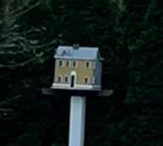 Modell av et lite to-etasjers hus med hvit base og mørkt tak, montert på en stang foran trær