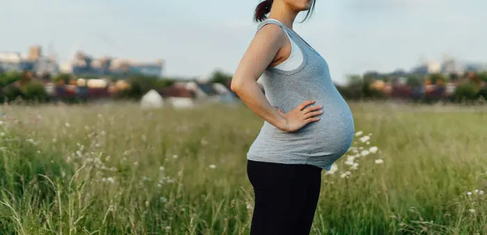 Gravid kvinne står selvsikkert i et felt med en byhorisont i bakgrunnen, iført en ermeløs topp og leggings