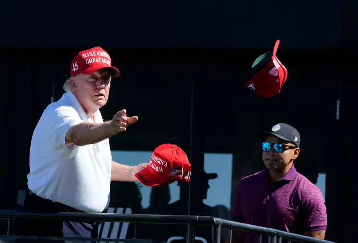 Donald Trump i en poloskjorte kaster en «Make America Great Again»; caps mens en annen person står i nærheten med solbriller og golfskjorte