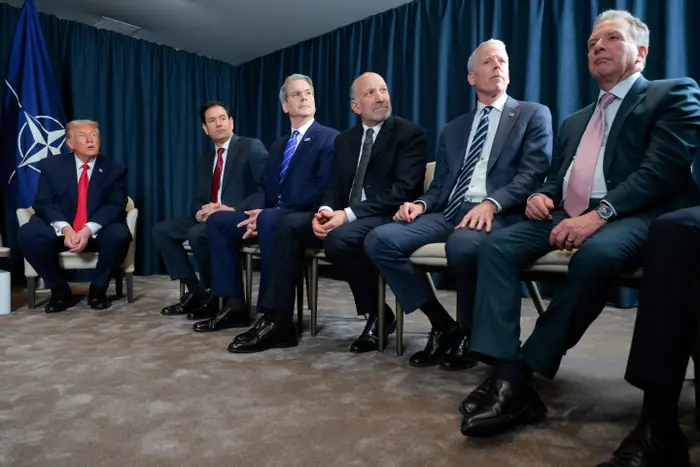 En gruppe på seks menn i dress sitter på rekke og rad og har en diskusjon foran et NATO-flagg