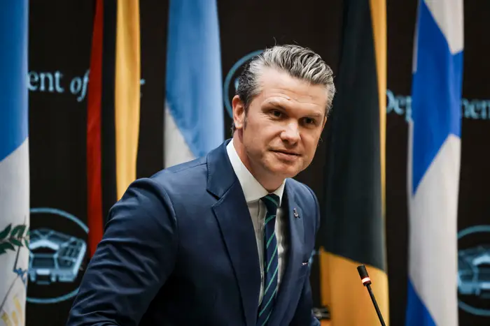 Pete Hegseth i dress taler ved et podium foran internasjonale flagg
