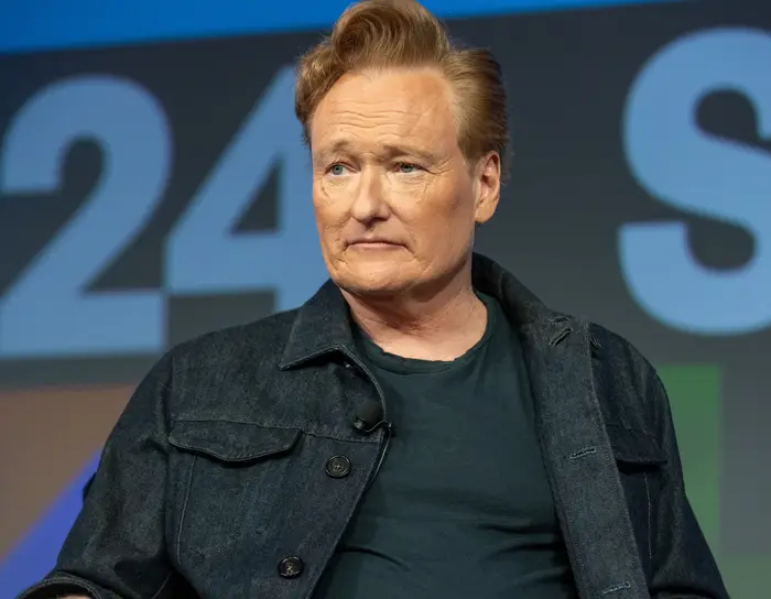 Conan O'Brien sitter foran et mønstret bakteppe, iført en dongerijakke over en T-skjorte, og ser til siden
