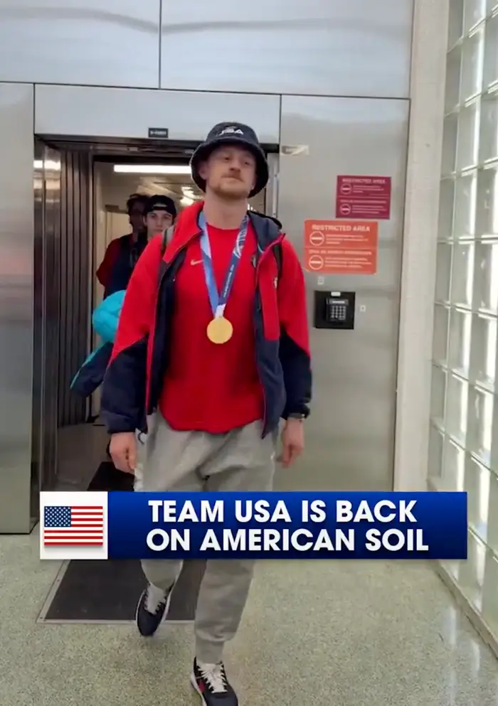 En person som har på seg en bøttehatt og atletisk klær, med en gullmedalje, går gjennom en flyplass. Teksten lyder: "Team USA er tilbake på amerikansk jord."