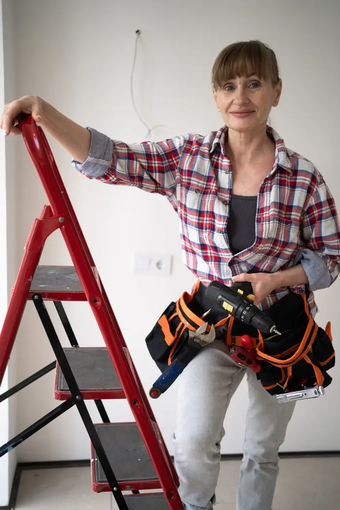 Person i rutete skjorte som holder et verktøybelte, står ved siden av en stige i et rom under oppussing