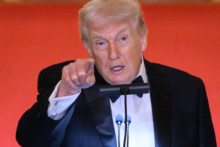 Mann i smoking snakker lidenskapelig ved et podium, gestikulerer fremover, mot rød bakgrunn