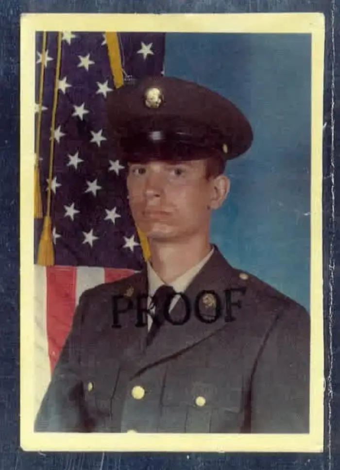 En person i militæruniform poserer formelt foran et amerikansk flagg med "BEVIS" stemplet over bildet