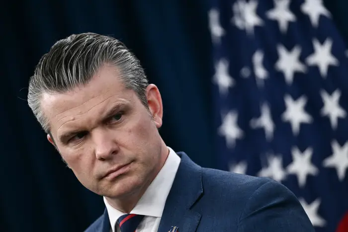 Pete Hegseth i dress, ser til siden, stående foran et amerikansk flagg som bakgrunn