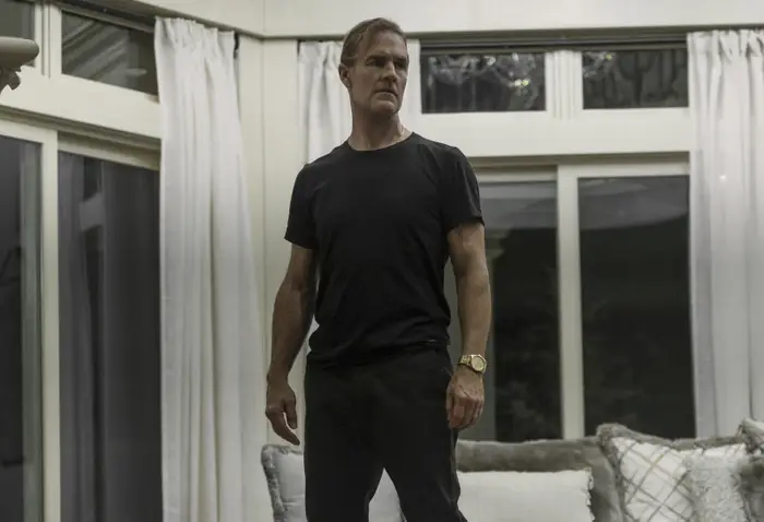 En person står selvsikkert i en stue, iført en uformell T-skjorte og bukser, og stirrer fremover med et alvorlig uttrykk