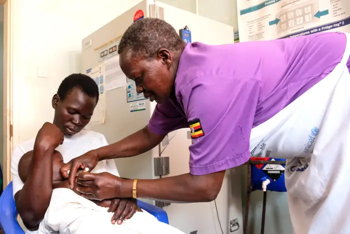 Helsepersonell som gir en vaksine til et barn som holdes av en forelder. Et medisinsk kjøleskap og informasjonsplakat er i bakgrunnen