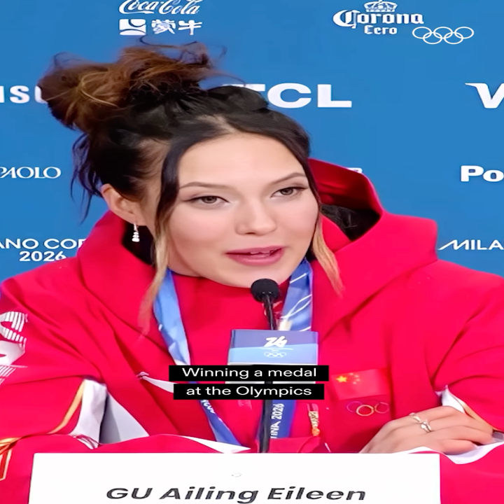 Person som snakker på en pressekonferanse, iført rød hettegenser og medalje, med tekstoverlegg: «Vinner en medalje i OL».