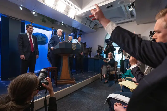 Politiker på podiet under pressebriefing, journalister sittende og rekker opp hendene, kameraer som fanger scenen