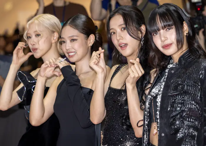 Fire kvinner poserer på en rød løper og gjør fingerhjertebevegelser. De har på seg elegante, stilige svarte antrekk, med fotografer i bakgrunnen