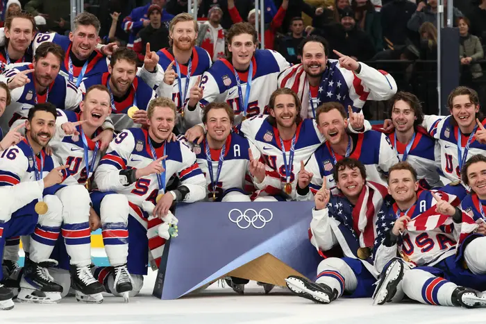 Team USA herrehockey feirer OL-seieren, alle iført medaljer og lagtrøyer, noen drapert i amerikanske flagg, på isen med et trofé