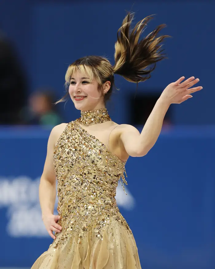 Kunstløper opptrer på is, smiler, med hår i bevegelse, iført en paljett, asymmetrisk kjole med høy halsdesign