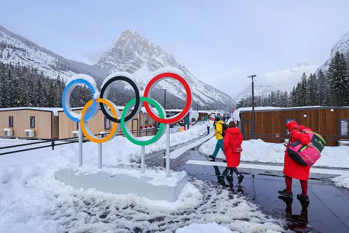 Folk går på en snødekt sti langs store olympiske ringer i vinterlige fjellomgivelser, omgitt av hytter og snødekte trær