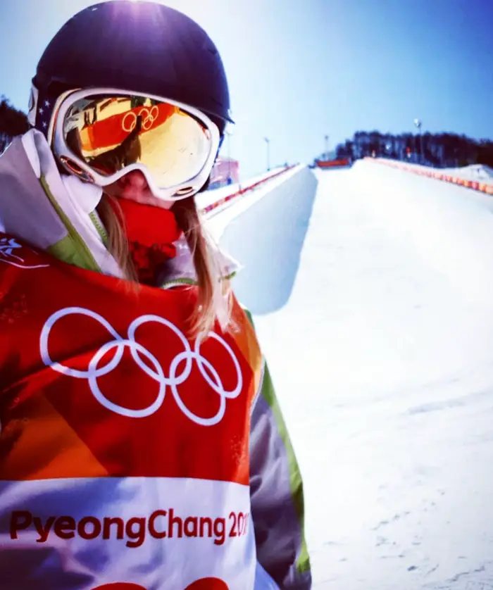 En idrettsutøver på en snødekt skibane iført olympisk utstyr og hjelm, med "PyeongChang 2018" synlig på klærne deres