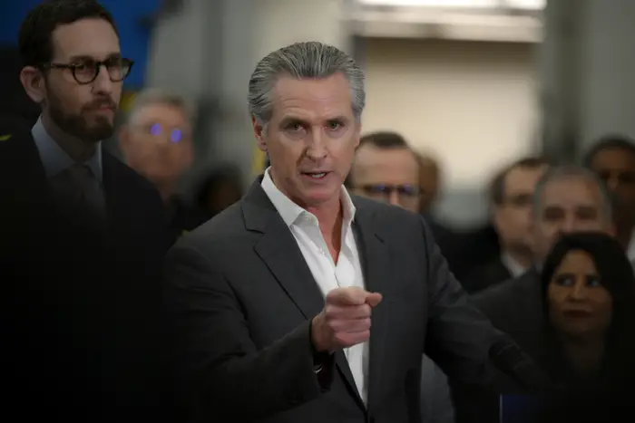 En person i dress snakker ved et podium, peker fremover, omgitt av en gruppe mennesker, foreslår en pressekonferanse eller offentlig kunngjøring