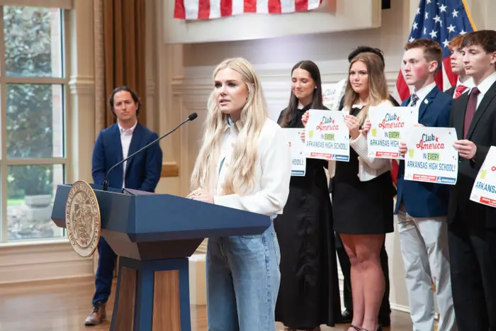 En kvinne snakker ved et podium, omgitt av unge voksne som holder skilt. Et amerikansk flagg er i bakgrunnen