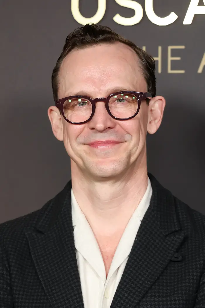 Person som smiler til arrangementet, har på seg briller og en blazer med mørk tekstur over en lys skjorte. Nøytralt bakteppe med delvis synlig eventskilt