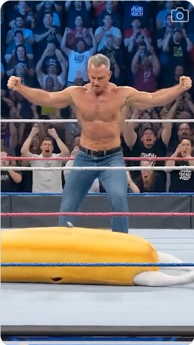 En muskuløs bryter i jeans poserer triumferende i ringen, med en oppblåsbar gjenstand ved siden av seg. Tilskuere jubler fra tribunen