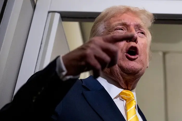 En person gestikulerer mens han snakker animert, iført en dress med et lyst slips. Uttrykket antyder selvsikkerhet eller at det haster