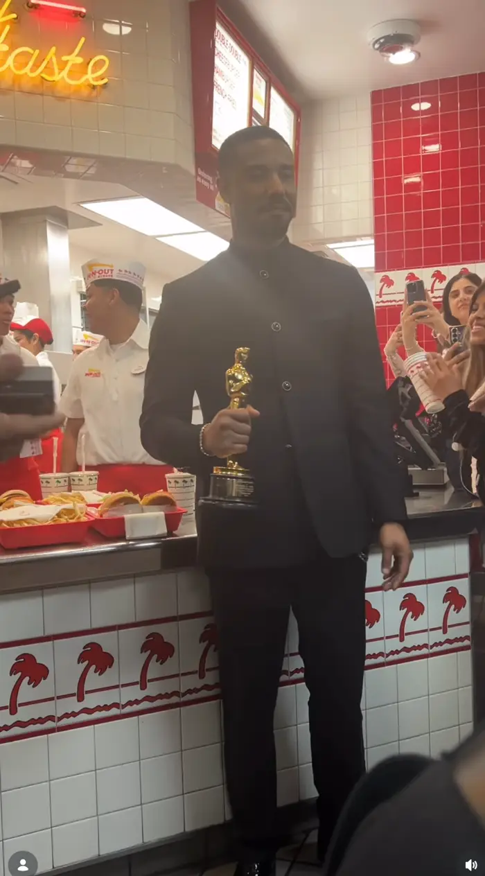 Person i mørk dress holder en pris inne på en gatekjøkkenrestaurant, med ansatte og kunder rundt, noen tar bilder