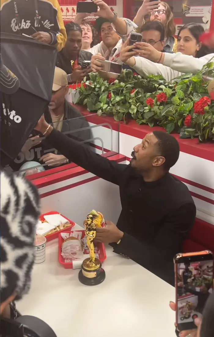 Mann i svart dress holder et trofé mens han sitter på en gatekjøkkenrestaurant. Fans og fotografer omgir ham og fanger øyeblikket