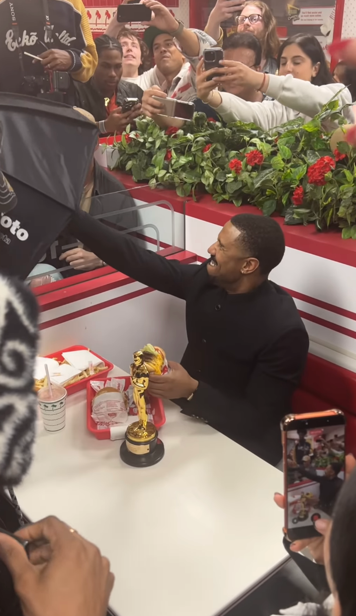 Person i formelle antrekk som holder gullstatue ved en fastfoodbod, omgitt av fans som tar bilder