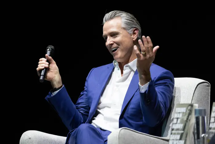 Person i dress sitter på en stol, snakker i en mikrofon, gestikulerer med én hånd under et arrangement