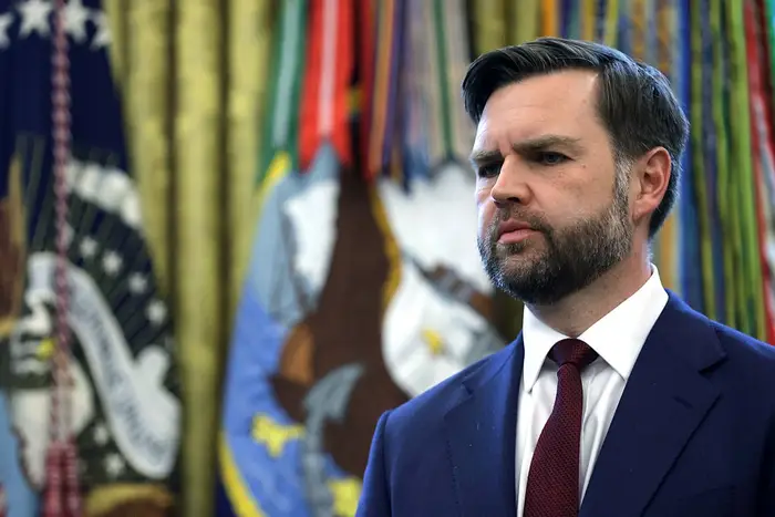 Mann i dress med et seriøst uttrykk står foran flagg med emblemdesign