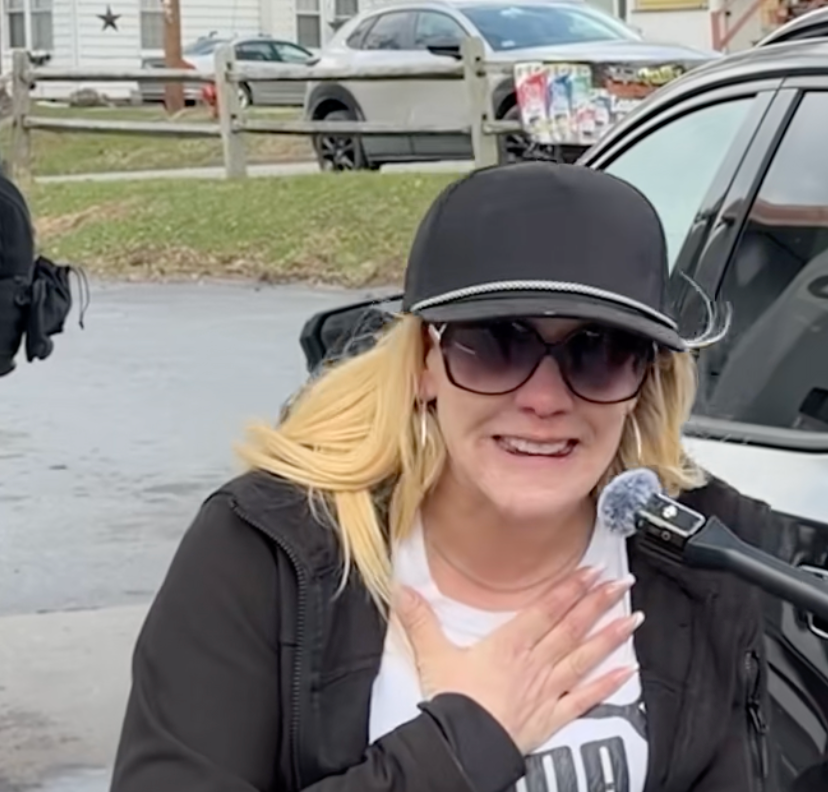 Person som står utenfor og ser emosjonell ut, har på seg solbriller og en caps, og holder en mikrofon nær munnen