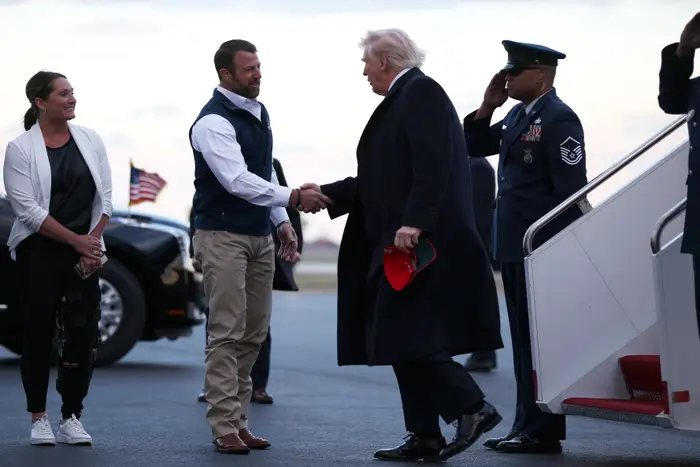 En mann i dress og frakk håndhilser på en annen mann i uformelt antrekk ved siden av en flytrapp. En uniformert offiser hilser i nærheten
