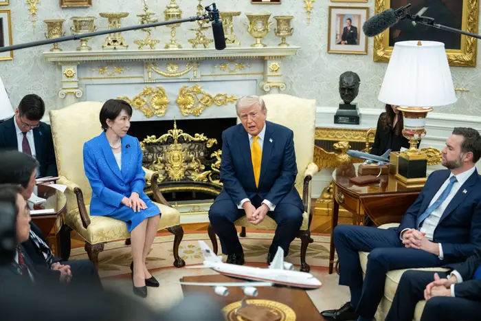 En gruppe tjenestemenn, inkludert en mann som snakker og en kvinne i dress, sitter i et formelt rom med utsmykket dekor og modellfly utstilt