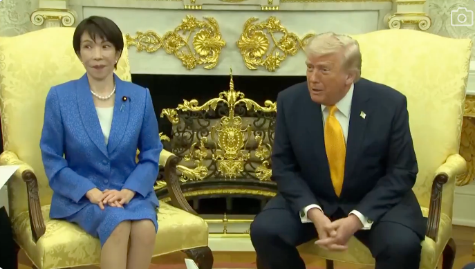 To personer sitter i overdådige stoler i en formell setting. Personen til venstre har på seg en klassisk dress, og personen til høyre har på seg en dress med et livlig slips